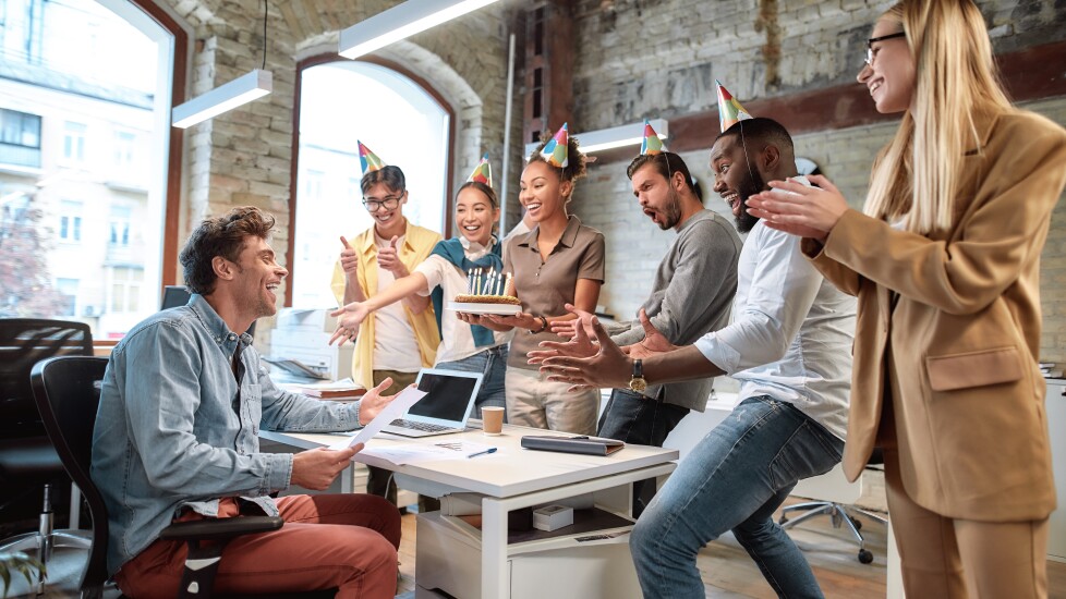 Office birthday celebration; employees clapping, woman holding birthday cake, surprising coworker