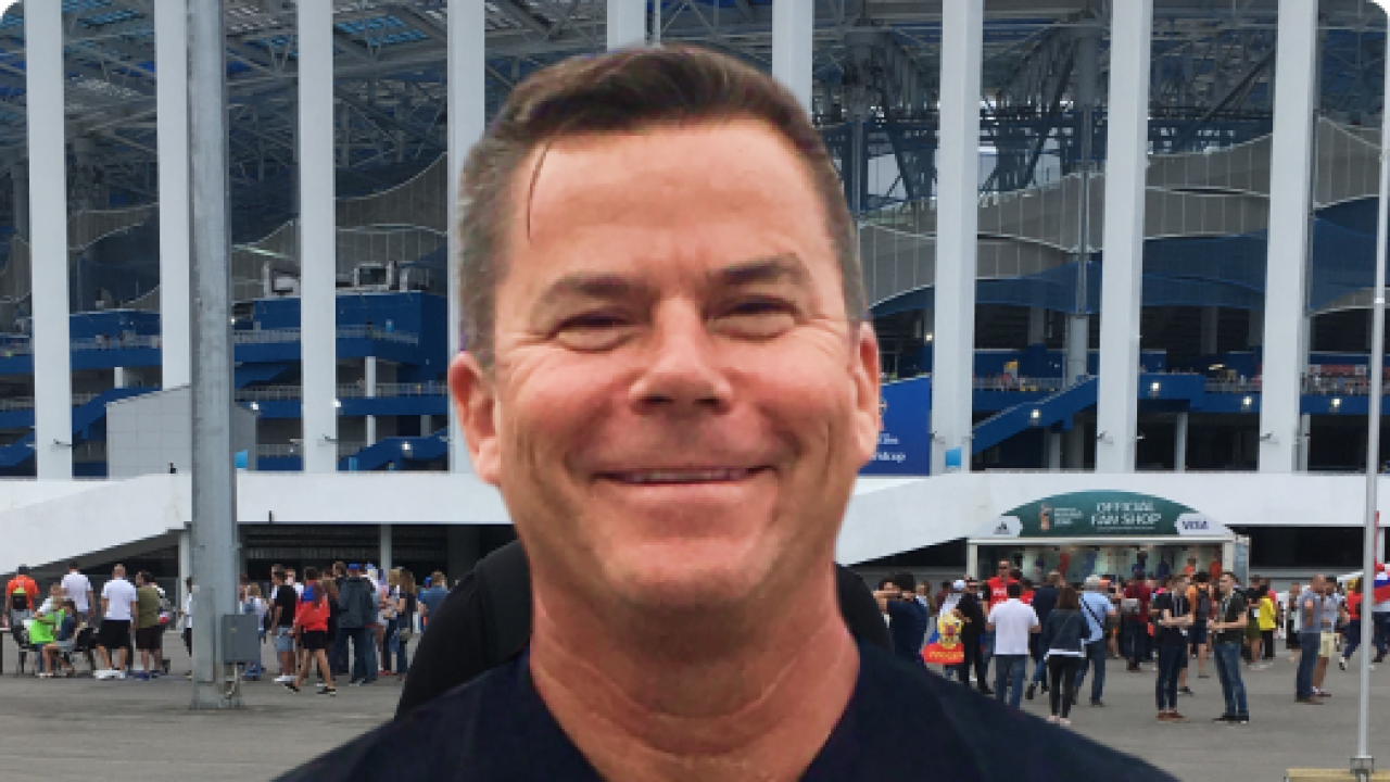 Man with brown hair and black collard shirt stands outside of a stadium entrance.
