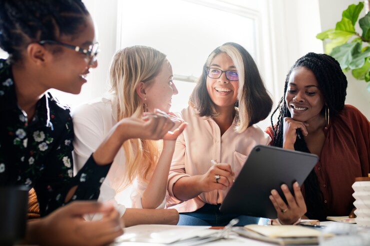 Four women gather around an iPad, smiling; they are backlit by sunlight streaming through a window.