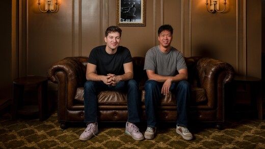 Two men sit on a brown leather couch in a dimly lit room with wall sconces and a framed photo of a pianist behind them.