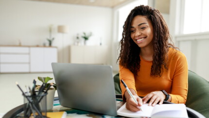Black woman sitting at desk, using pc writing in notebook