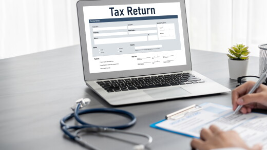 Tax return on laptop screen with stethoscope and person writing on clipboard