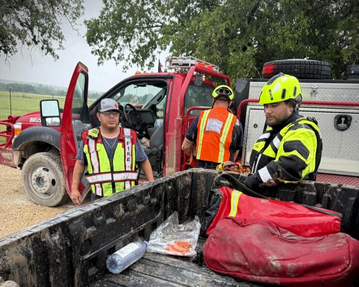 Three firefighters grab supplies from two mud-covered trucks in the rain to assist with Central Texas flood relief efforts