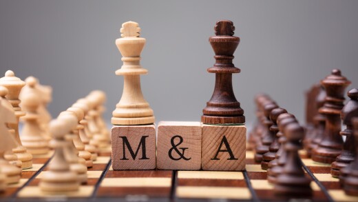 Wooden chess pieces in light brown and dark brown are lined up on a chessboard with the two king pieces sitting on top of blocks reading "M&A"