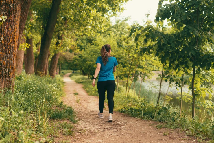 A woman dressed in a blue t-shirt and leggings is walking down a dirt path surrounded by trees and underbrush.
