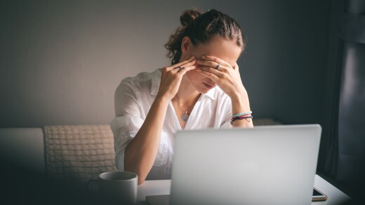 A young woman in a white shirt sits at a table in front of a laptop.