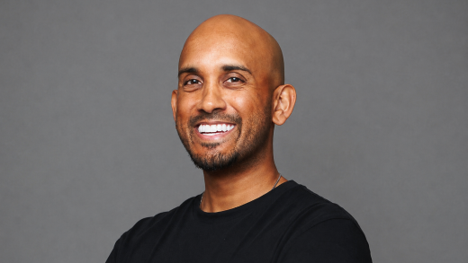 Smiling man with black shirt standing against a gray background.