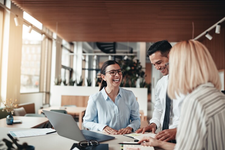 Three people in an office working at a table with a laptop and notebooks on the surface.