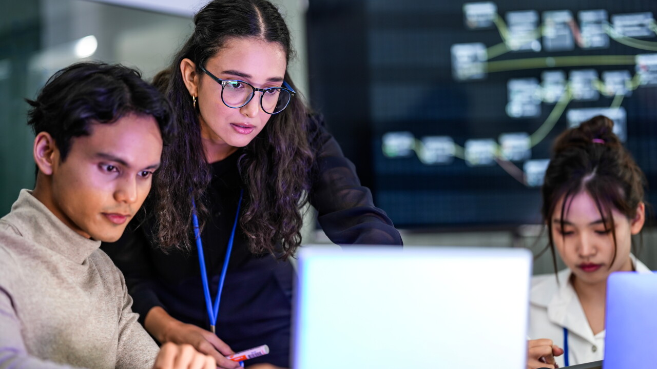 A diverse group of professionals in a corporate workshop on AI development. A female instructor supervises as the team learns to code advanced machine learning models on their laptops.