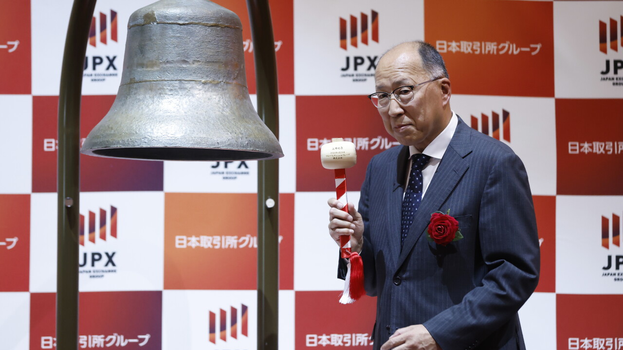 Man in suit holding a mallet next to a large bell in front of a red and white JPX (Japan Exchange Group) backdrop.