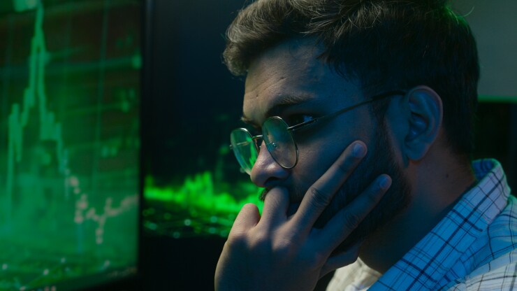 A man wearing glasses looks intently at a screen displaying green financial data and stock charts, suggesting focus on cryptocurrency or digital asset analysis.