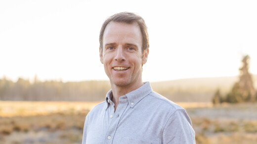A smiling man in a light grey button-down shirt outdoors in golden hour sunlight with a blurred landscape background.