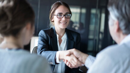 A young woman wearing glasses and a suit shakes hands with an older man.