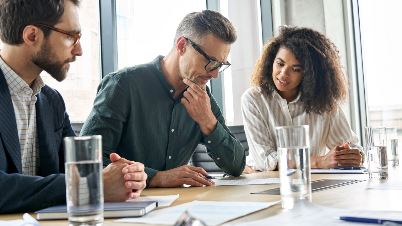Three corporate employees sitting at a conference stare down at a piece of paper, looking concerned.