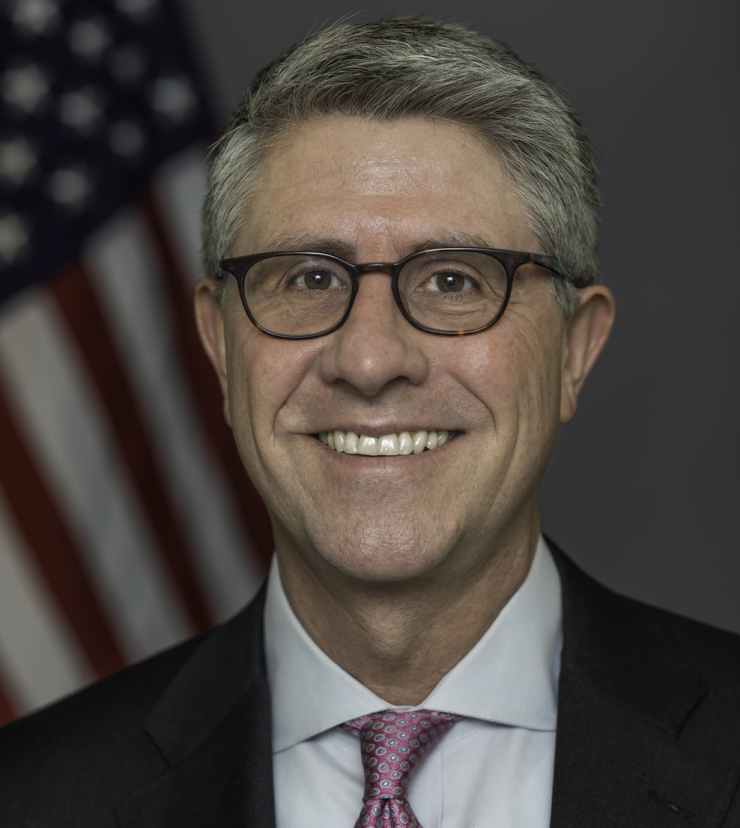 Headshot of man with gray hair and glasses wearing a suit and patterned pink tie standing in front of a blurred American flag in the background.