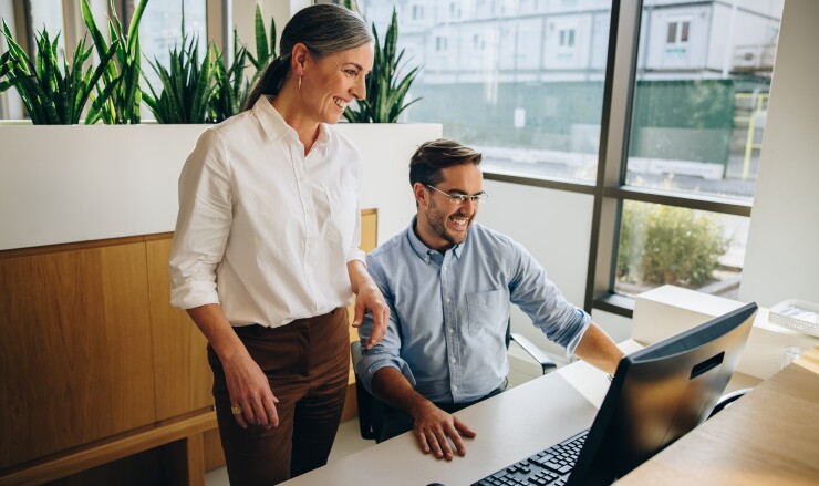 Two people, one standing and the other sitting, while they look at a computer monitor.