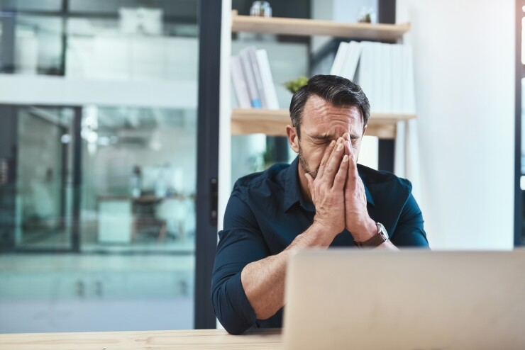 A man puts his face in his hands in frustration. He's at his desk in front of his laptop.