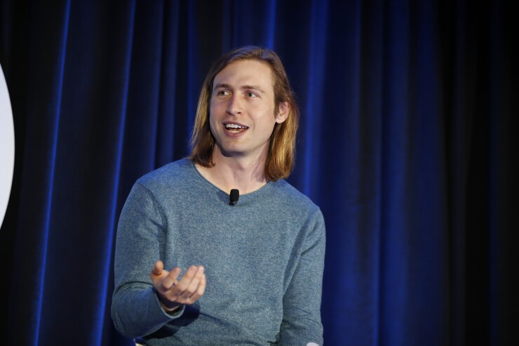 Man with long blond hair and blue tee speaks on a stage with blue curtains in the background.