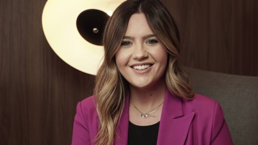 Smiling woman with wavy blonde-highlighted hair wearing a magenta blazer, sitting in front of a modern circular light.