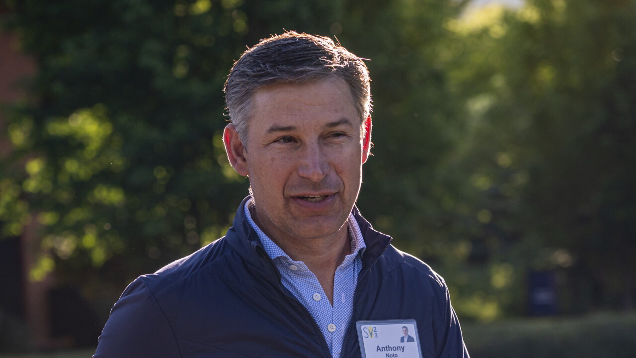 Anthony Noto wearing a blue vest and name tag, standing outdoors with golden sunlight highlighting his hair.