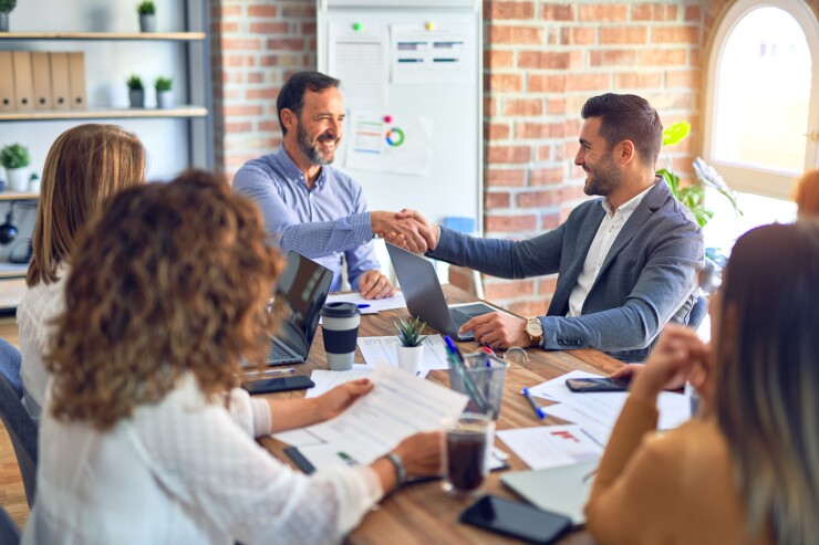 Group of employees sitting around table in meeting; two male employees shaking hands