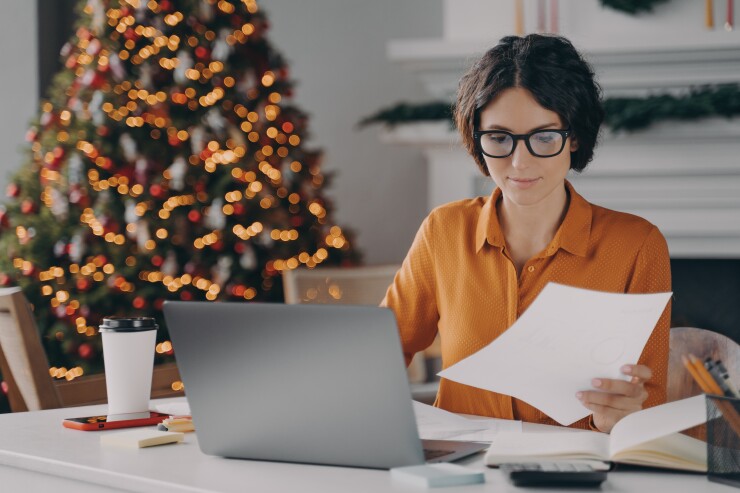 A woman works at her desk in what looks like a living room with a fireplace; a Christmas tree is behind her.