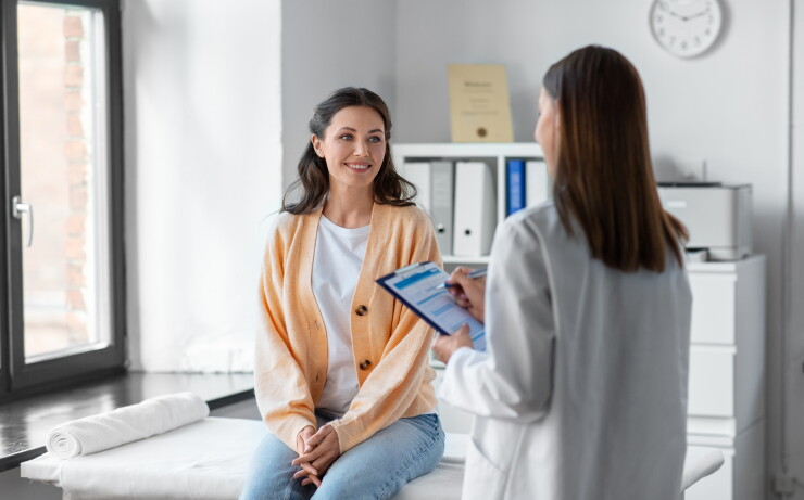A woman in an orange cardigan sits on an examining table while a doctor with a clipboard asks her questions