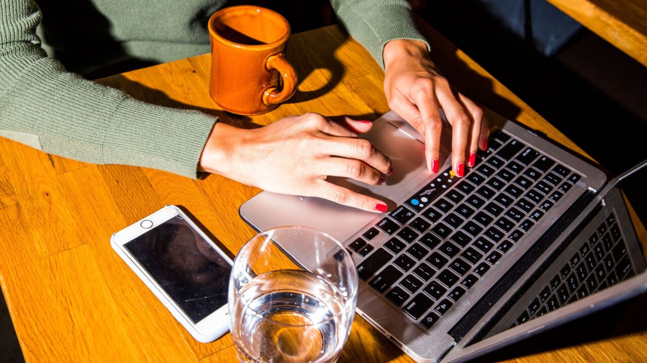 Woman with red manicure typing on a laptop with a glass of water on her desk.
