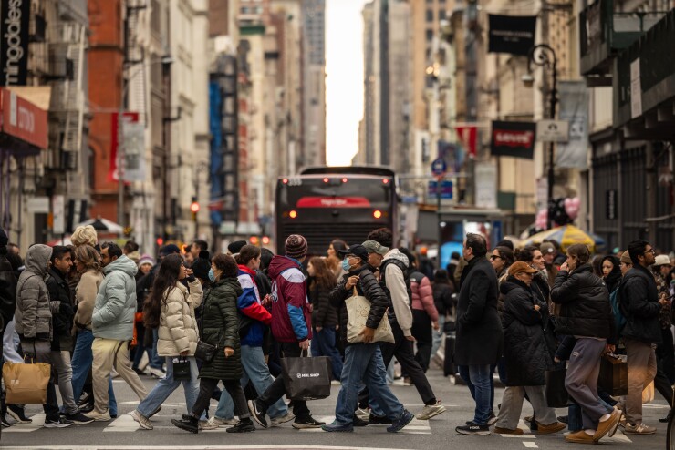 Pedestrians crossing a busy city street, some holding shopping bags, with a bus in the background