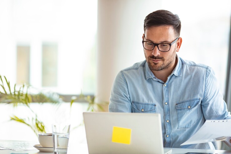 Man with glasses sitting at table desk working on laptop paper in hand