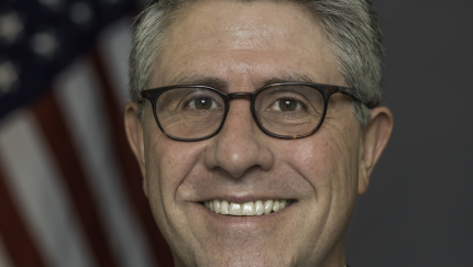 Headshot of man with gray hair and glasses wearing a suit and patterned pink tie standing in front of a blurred American flag in the background.