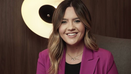 Smiling woman with wavy blonde-highlighted hair wearing a magenta blazer, sitting in front of a modern circular light.
