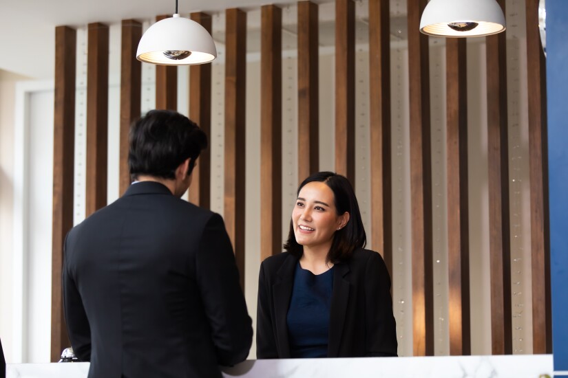 A hotel front desk employee, a woman in a blue top and black blazer, is talking to a man in a black blazer.