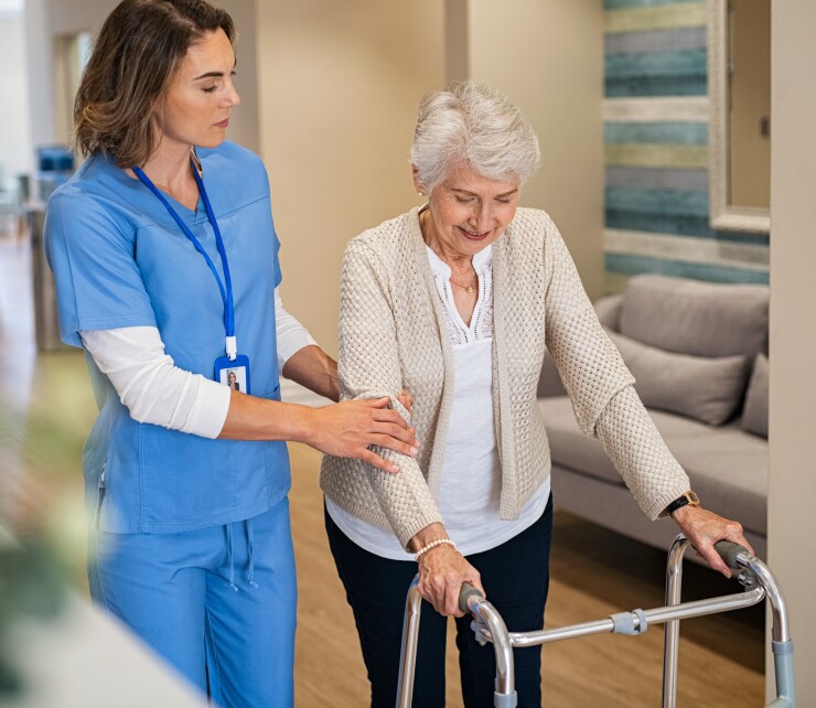 Stock photo of an elderly person with using a walker with assistance from a woman wearing medical scrubs