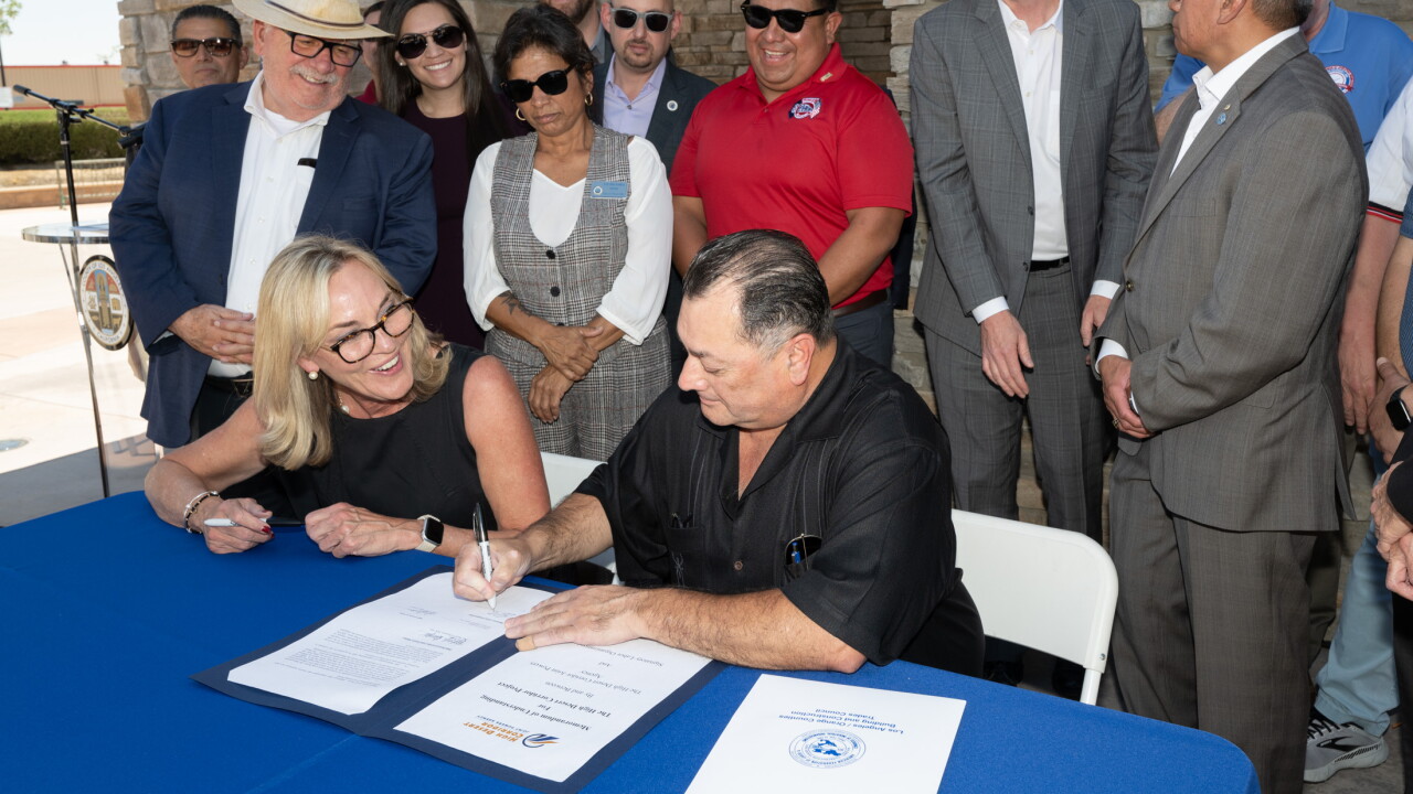 Two people at a table signing documents with at least nine people standing behind them