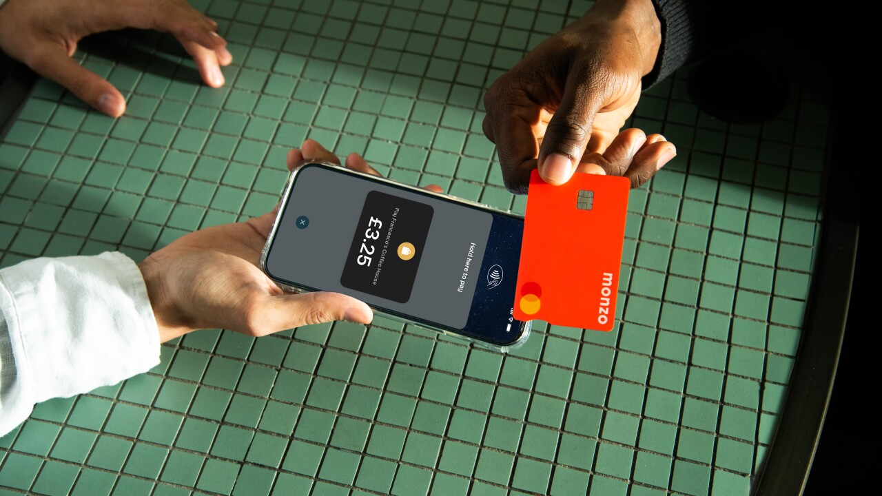 A close-up photograph shows a contactless payment being made. A person with a light-skinned hand holds a smartphone displaying a Monzo payment screen for £3.25 at "Francesco's Coffee House," with the text "Hold here to pay." Another person with a dark-skinned hand holds a bright red Monzo debit card just above the phone. The hands and devices are over a green, square-tiled table surface under bright, directional light.