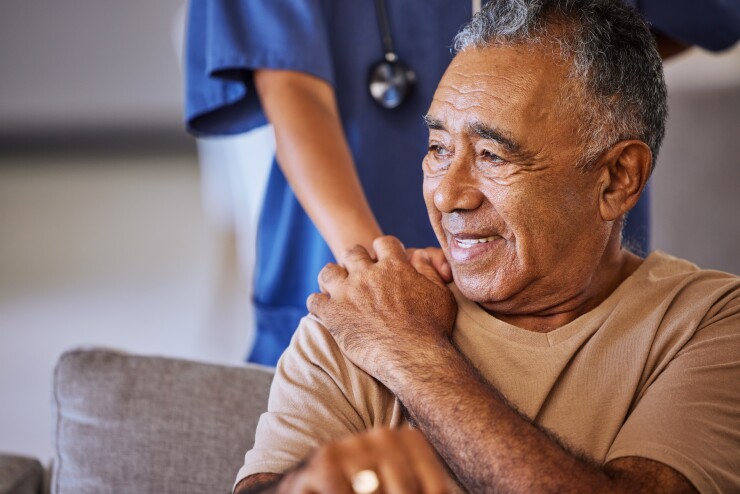 A nurse is holding an elderly man's shoulder; the elderly man touches her hand back.
