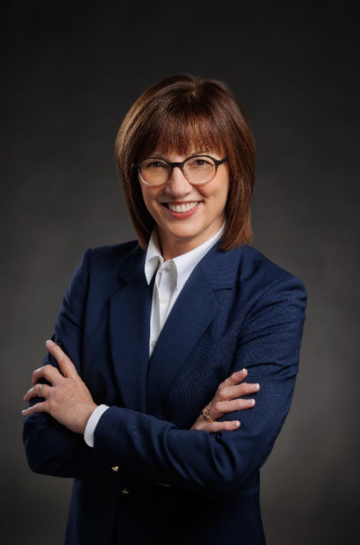 Kelly Davids standing with her arms crossed, smiling, wearing a button-down shirt and a blue blazer.