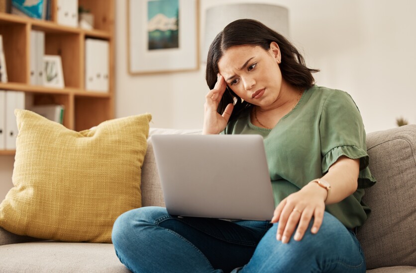 A woman is sitting on her gray couch with her laptop in her lap, looking confused; a yellow pillow is next to her, and it's daytime.