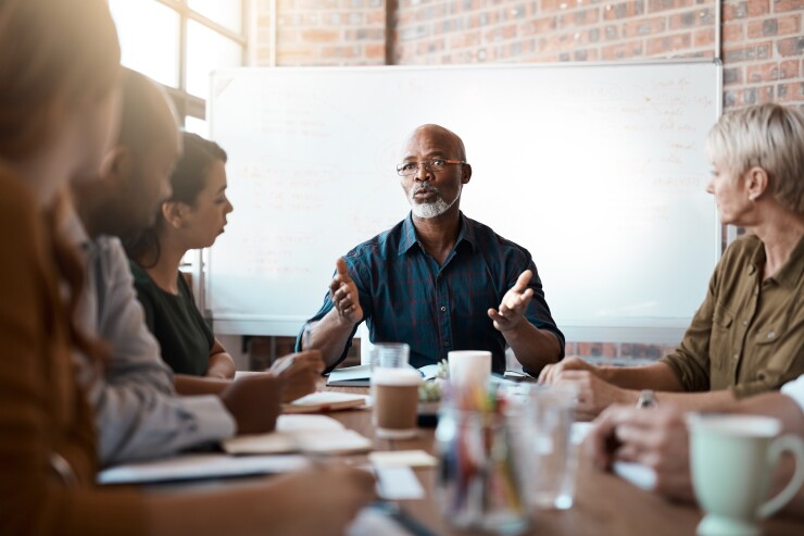 A Black man at the head of a conference table is explaining something to a group of colleagues.