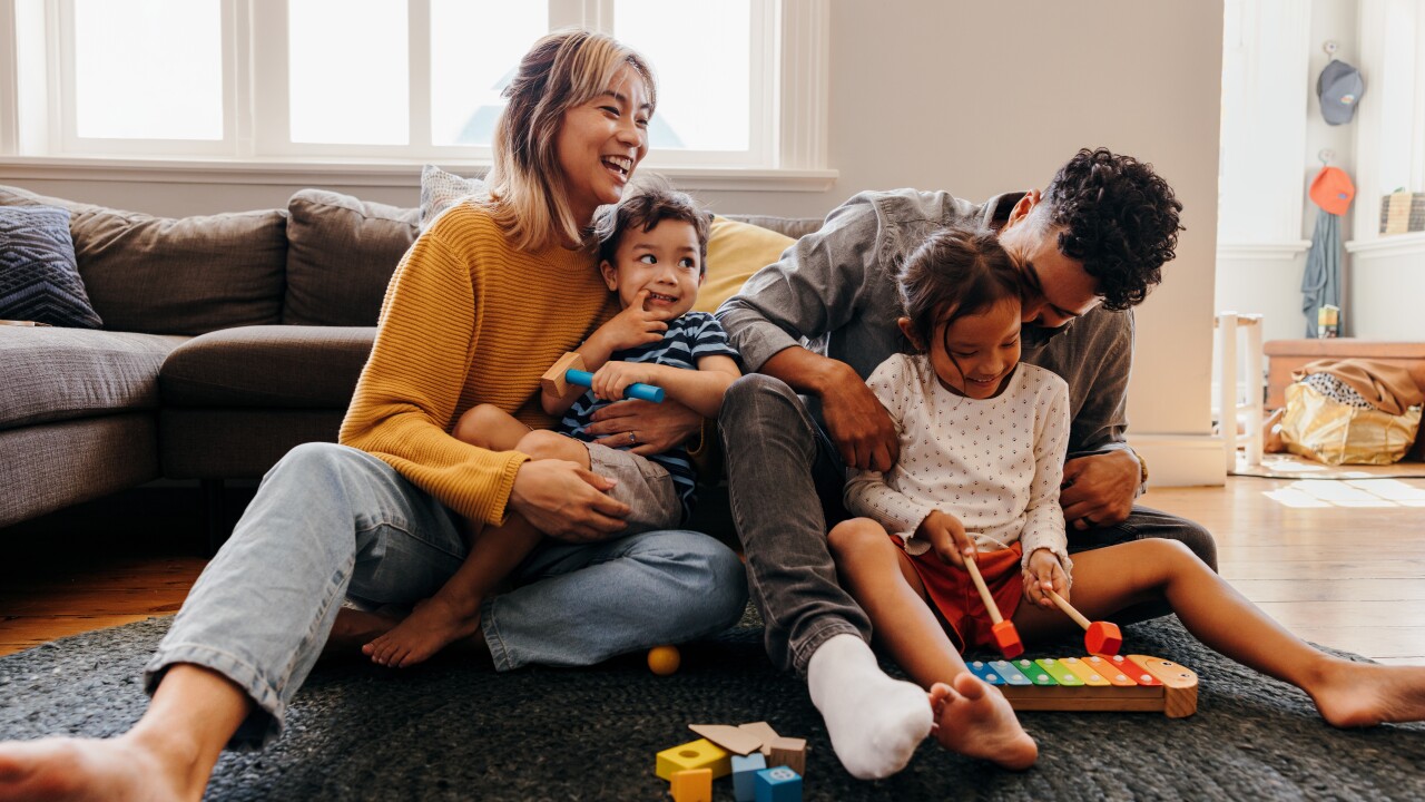Parents with a son and daughter are holding them in their laps while laughing in their living room; there are colorful toys on the rug at their feet.