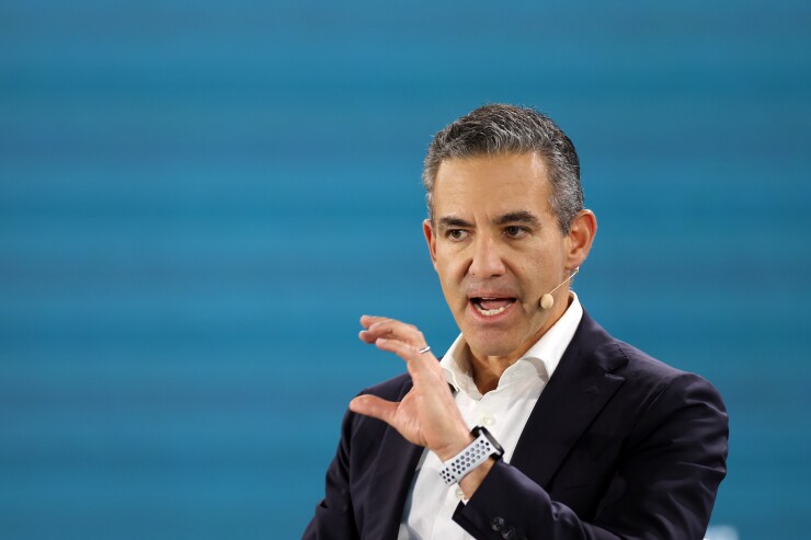 A man in a navy blazer and white shirt speaks with hand gestures and a headset microphone against a blue background.