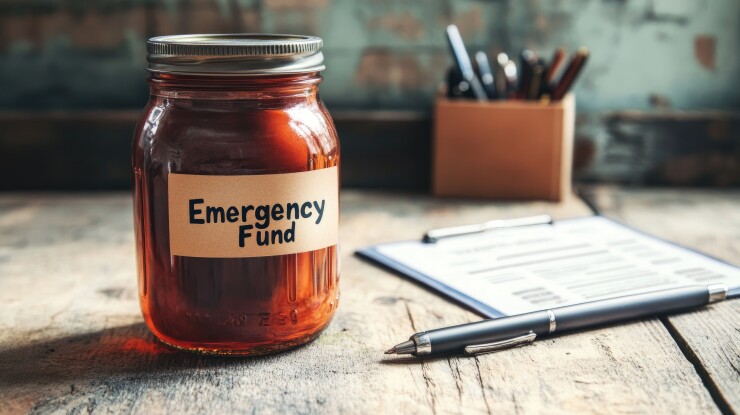 A jar with a label that says "Emergency Fund" sits on a table next to a pad and pen.