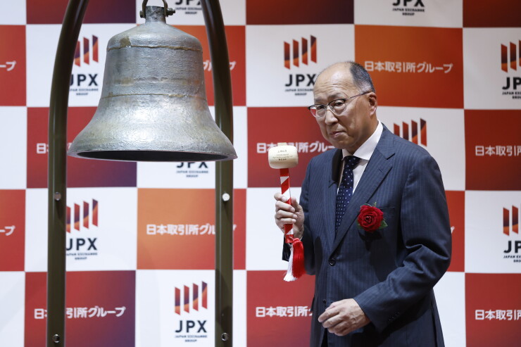 Man in suit holding a mallet next to a large bell in front of a red and white JPX (Japan Exchange Group) backdrop.