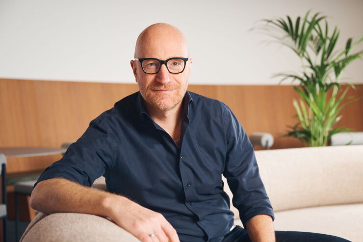 Bald man wearing black glasses and a blue collared shirt sits on beige couch with tall green plant in background.
