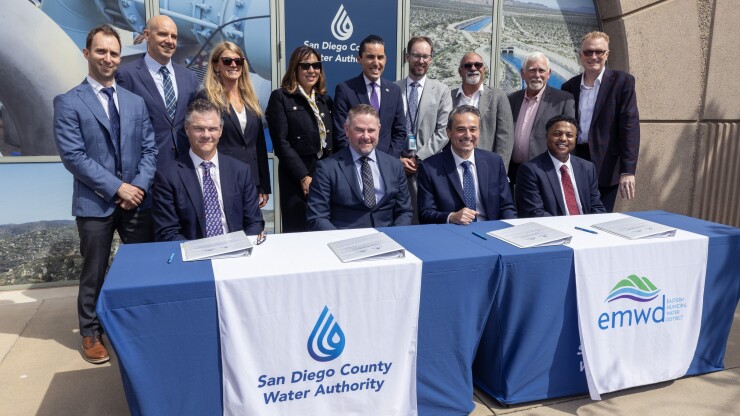Four men in suits at table labeled for San Diego County Water Authority and Eastern Municipal Water District with nine people standing behind them.