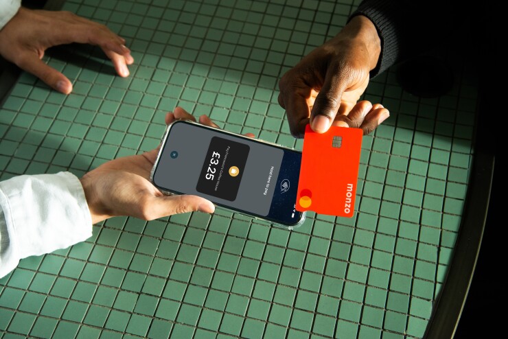 A close-up photograph shows a contactless payment being made. A person with a light-skinned hand holds a smartphone displaying a Monzo payment screen for £3.25 at "Francesco's Coffee House," with the text "Hold here to pay." Another person with a dark-skinned hand holds a bright red Monzo debit card just above the phone. The hands and devices are over a green, square-tiled table surface under bright, directional light.