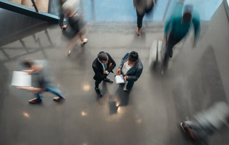Two people standing in the lobby of an office looking at a tablet while blurred people walk past.
