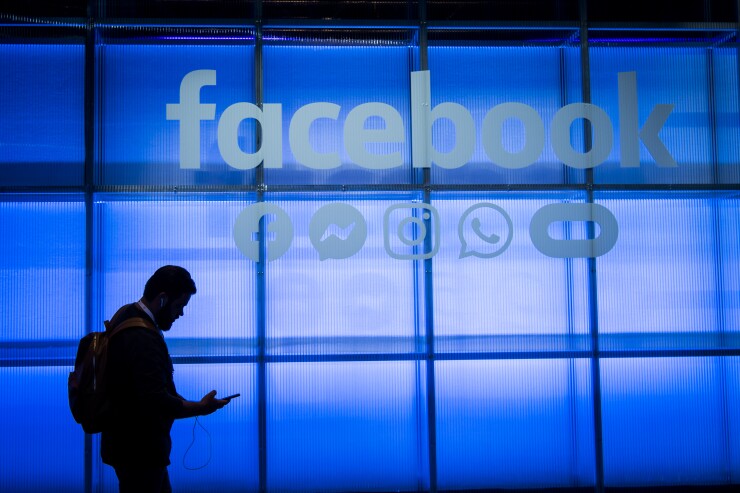 An attendee looks at a mobile phone while walking under a Facebook logo in the demonstration room during the F8 Developers Conference in San Jose, California, on April 30, 2019.