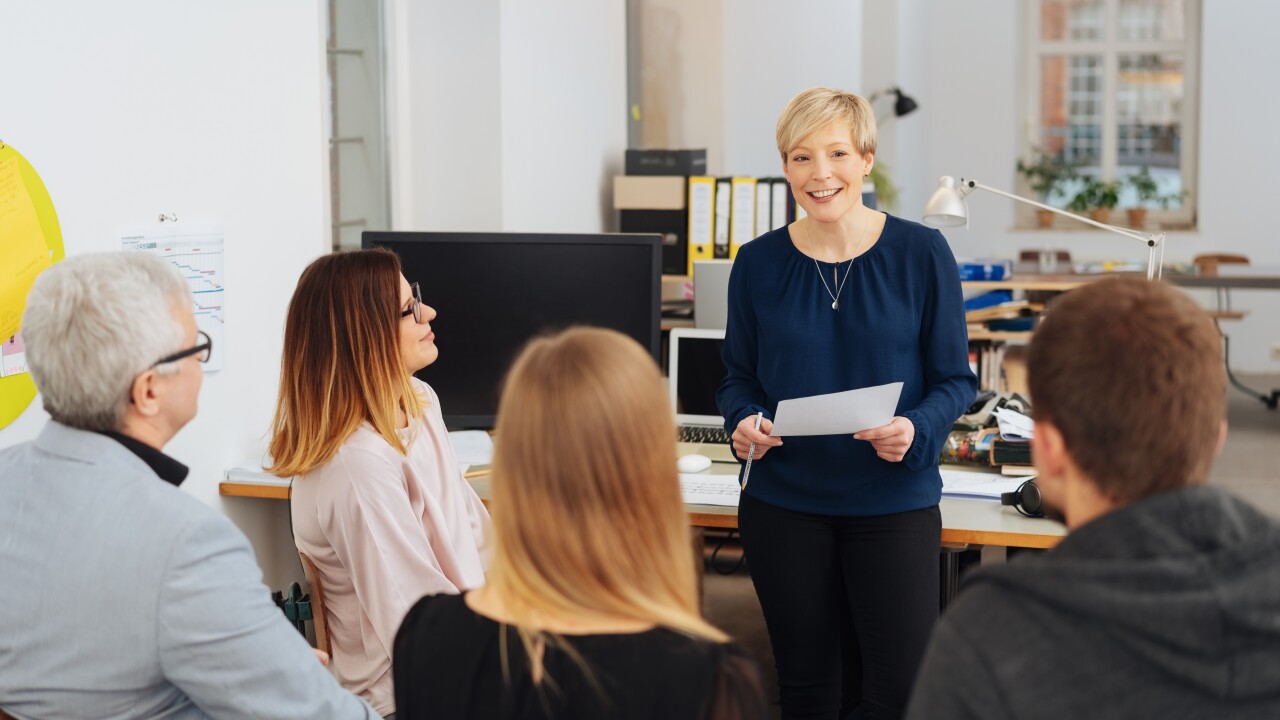 Group of seated employees listening to female employee who is smiling, leaning on desk